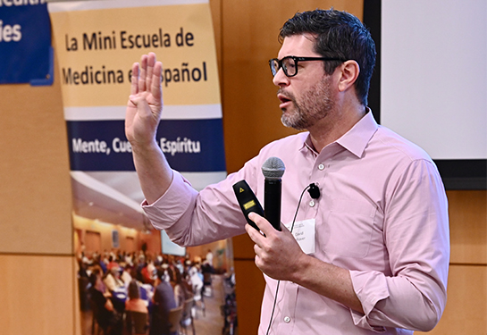 A man in glasses and pink shirt holds microphone in left hand and gestures with his right, while addressing a conference 