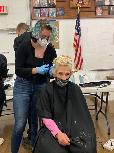 A woman seated with a black beauty salon cape gets her hair cut from a hairdresser standing behind her