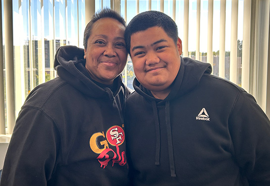 Woman with short hair and black sweatshirt stands next to young man with black sweatshirt.