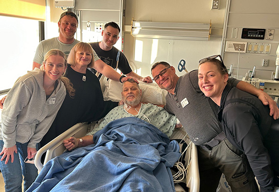 Six smiling family members stand around a hosptial bed where an older male patient rests