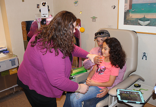 A child life specialist shows a father whose young daughter is sitting on his lap, how to hold her comfortably to receive a flu shot. 