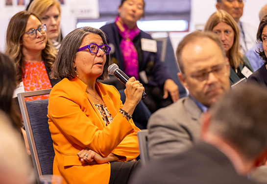 A woman in an orange suit jacket seated at a chair speaks into microphone conference room surrounded by several people