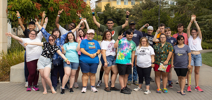 A group of 20 smiling college students gather on a sidewalk for a group photo.