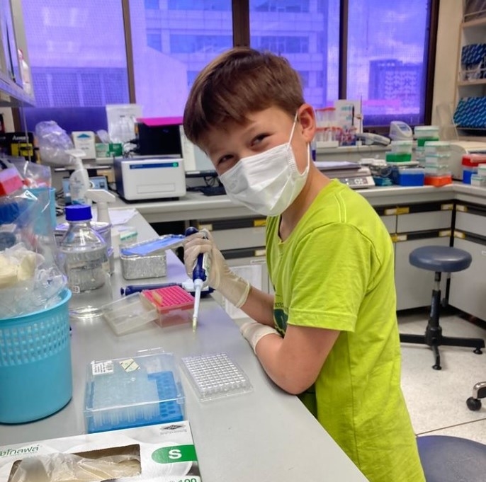 A young boy wearing a green t-shirt, surgical gloves and mask holds a dropper in a laboratory