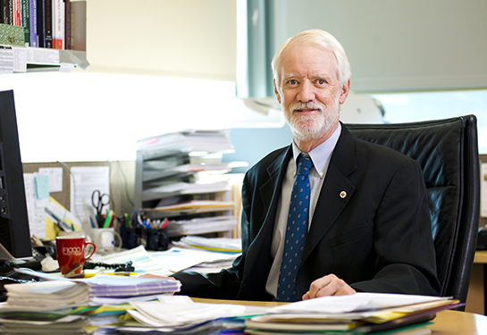 A man with whitish-gray hair sits behind a desk in an office. 