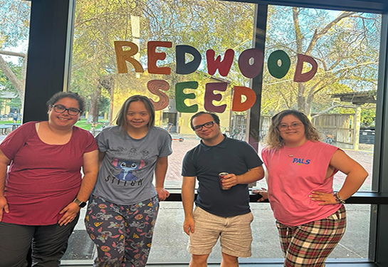 Four students stand before a large window with a sign that reads "Redwood SEED"