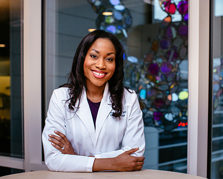 Woman in white coat sitting at table with arms cross with multi-colored glass sculpture hanging behind her.