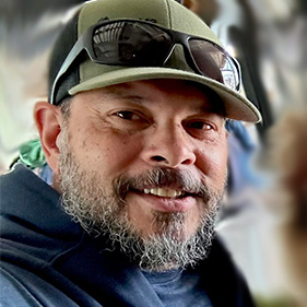 Headshot of a man with a beard and moustache in a blue shirt and brown hat.