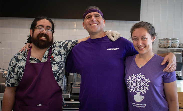 Three café employees stand together in front of a coffee machine with their arms around each other, smiling. 