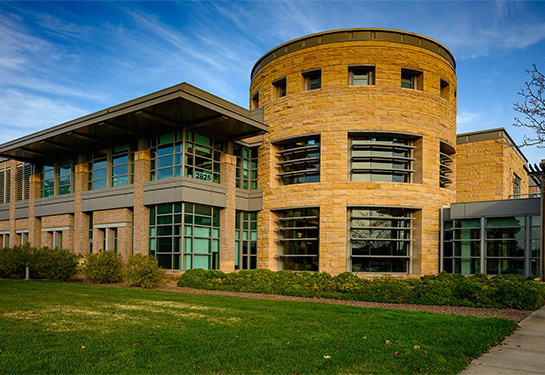 The UC Davis MIND Institute, a large sandstone and glass building surrounded by green bushes and concrete sidewalks. 