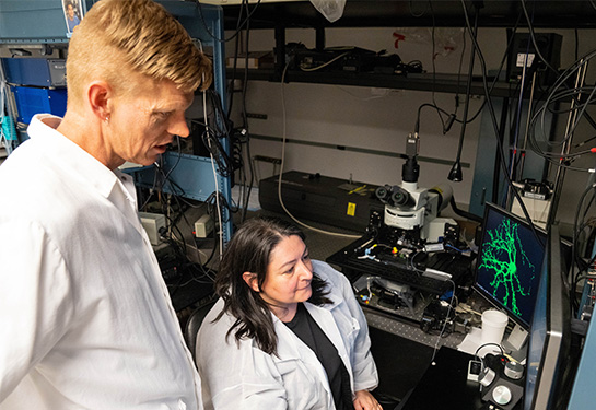 Two scientists in white lab coats in a research lab look at a computer monitor. 