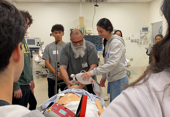 A man with a beard stands between high school students and teaches them to pump air into a training manikin&#x2019;s nose and mouth 