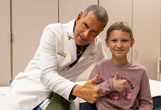 Male soctor wearing white coat and female patient sitting on an exam room bed giving a thumbs up