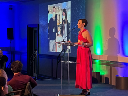 Woman with red dress at podium speaking with photo of child and two adults on screen behind her.