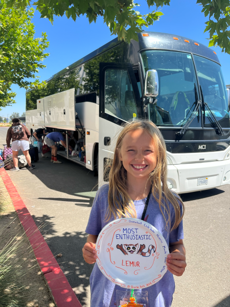 Blonde girl smiling with a paper plate sign that reads “Lemur” and “Enthusiastic” and standing in front of a bus.