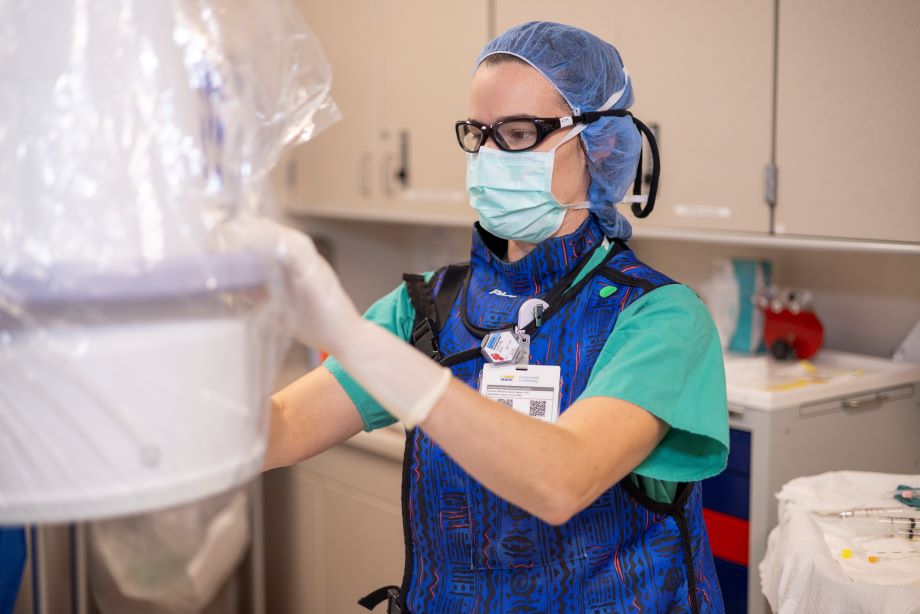 A doctor in scrubs, hairnet, goggles and mask positions x-ray equipment in a patient procedure room. 