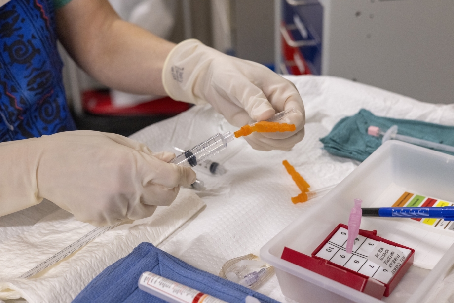 A gloved hand inserts a needle into a tube above a tray table that holds medical equipment for pain procedures.