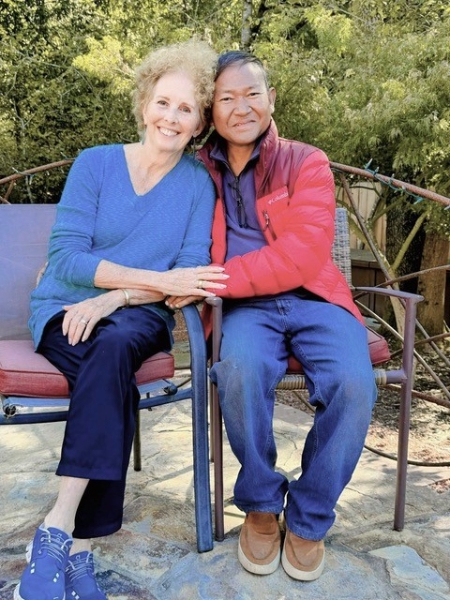 Cynthia McReynolds and Wee Simla sitting outside in metal chairs while leaning in towards each other and holding hands.