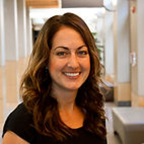 A woman with long brown hair, wearing a dark-colored shirt smiles in a hallway