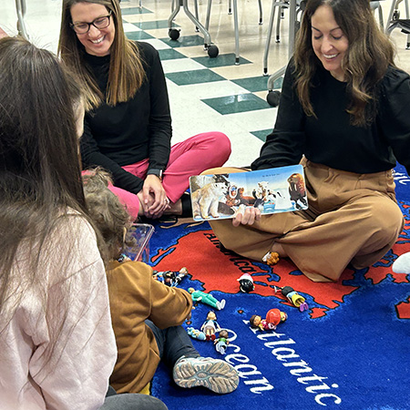 A woman sits on a rug with two other woman and a young girl, reading a picture book.