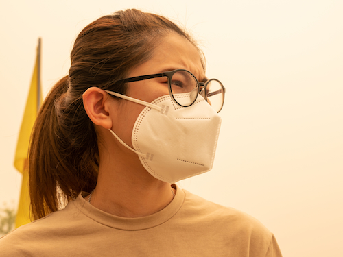 Worried-looking woman wears a mask against a hazy pink background of bad air.