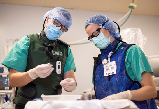 Two doctors in scrubs, masks, hair nets and protective vests stand over a tray table. One doctor holds a needle. 