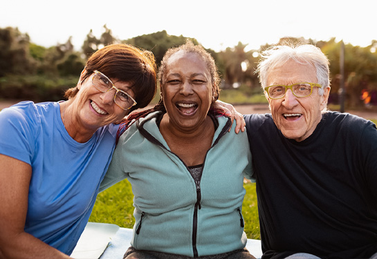 Three older adults sitting on grass next to each other smiling at camera