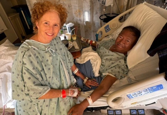 Wee Simla laying in hospital bed and Cynthia McReynolds wearing hospital gown sitting on edge of hospital bed.