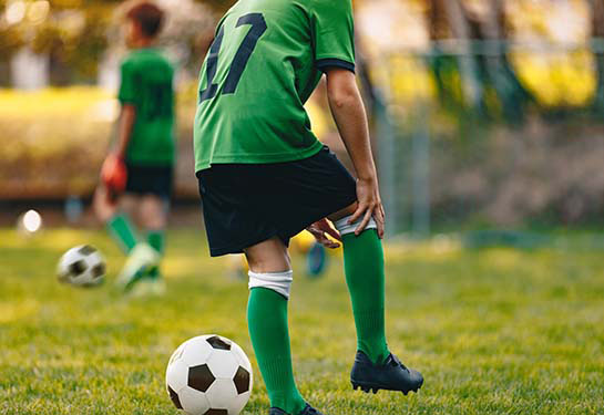 A boy wearing a green and black soccer uniform reaches down to hold his knee, a soccer ball next to him on a field.  