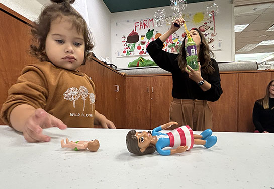 A  young girl plays with plastic toys at a table while a woman blows bubbles in the background