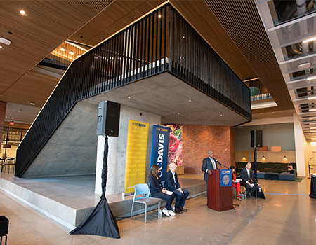 Standing at a lectern, UC Davis Chancellor Gary S. May speaks into a microphone as a seated man and woman look on