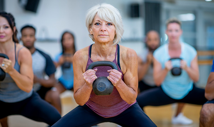 An older white woman doing a squat with a kettlebell, while participating in a co-ed, multi-ethnic, fitness class