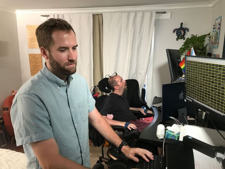 Dr. Nicholas Card works on a computer with monitor showing brain waves. Nearby, BCI BrainGate trial participant Casey Harrell looks at another screen.