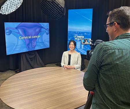 A woman sits behind a table for a television interview, with a TV screen to her right that says "cervical cancer" while a man behind the camera looks on.