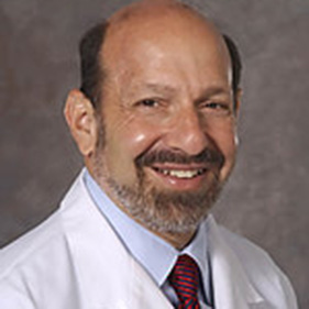 A physician in a white coat with a beard, smiles