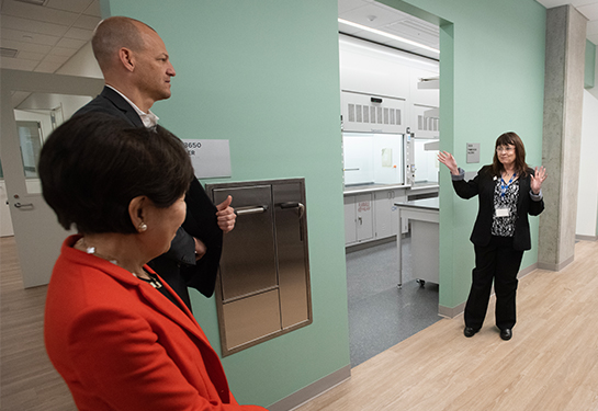 A woman, standing on right in a research laboratory, gestures with her hands in conversation with a man and a woman on left