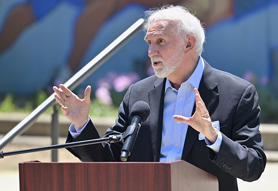 A man with a white beard in a navy blue suit jacket and blue collared dress shirt gestures with both hands at a podium outdoor