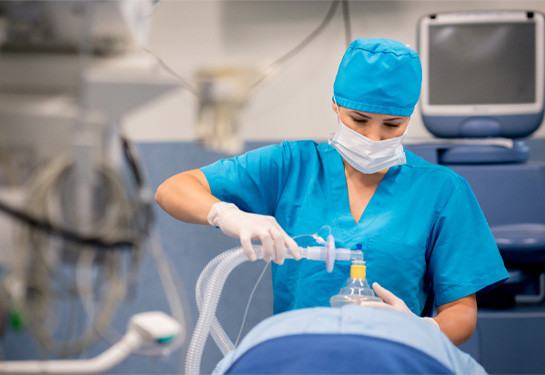 Nurse anesthetist in scrubs holding mask with tube over mouth of patient lying on the operating room table