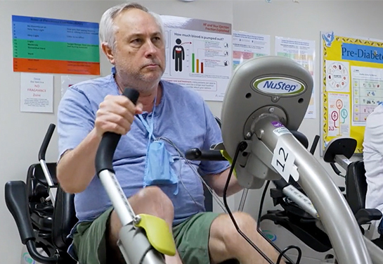 Paciente de la tercera edad, en camiseta y pantalones cortos, sentado sobre una máquina para hacer ejercicios de piernas y brazos