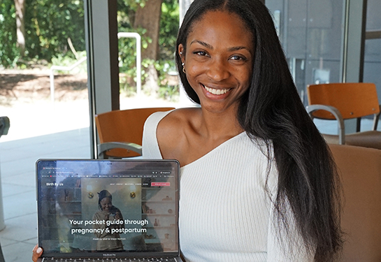 A woman wearing white sweater holds laptop with its screen that reads: &#x201c;Your pocket guide through pregnancy and postpartum&#x201d;