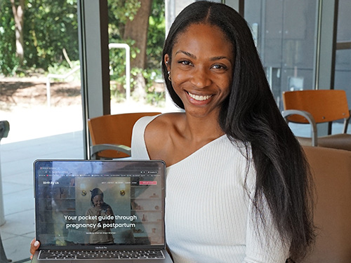 A woman wearing white sweater holds laptop with its screen that reads: “Your pocket guide through pregnancy and postpartum”