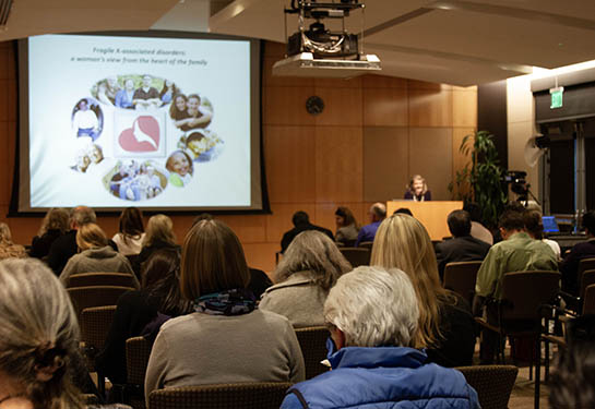 A crowd of seated people look toward a podium where a woman gives a lecture in an auditorium.