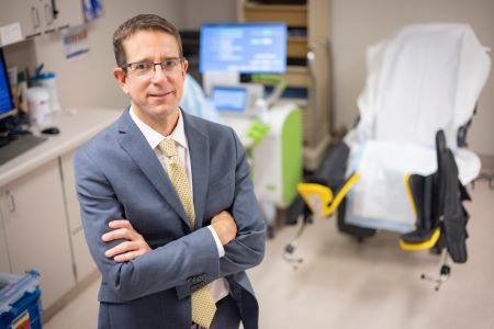 Man with light brown hair and suit standing in front of a patient chair in an exam room.