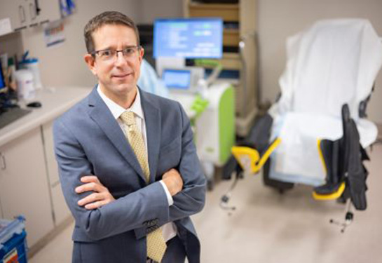 Man with light brown hair crossing arms and standing in patient exam room.