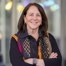 UC Davis School of Medicine Dean Susan Murin is smiling and standing in a brightly lit hallway with her arms crossed. She is wearing an orange, pink a