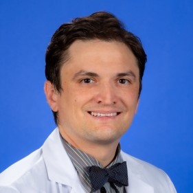 A portrait of a man against a blue background wearing a white coat, grey shirt and dark bowtie. 