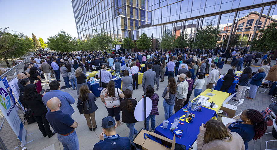 A large crowd gathers near a tall glass building