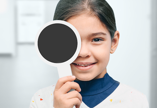 Niña con cabello recogido, sonriendo mientras se cubre un ojo con una herramienta oftálmica durante una evaluación de la vista.