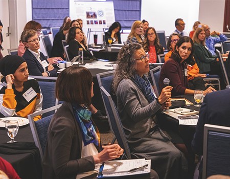 A woman seated at a table surrounded by several men and women holds a microphone to provide input to a panel of experts