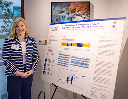 A woman with blond hair stands next to a research poster that highlights teamwork to support advance care planning in primary care